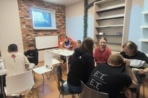 Grupa ludzi siedzi przy dwóch stołach. Pochylają się nad grami planszowymi. W tle duża półka na książki i pokój warsztatowy biblioteki.