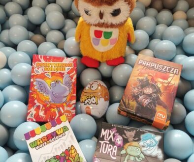 Pięć nowych gier planszowych. Ułożone na plastikowych kulkach w kolorze niebieskim. Za grami stoi kolorowa maskotka sowy.